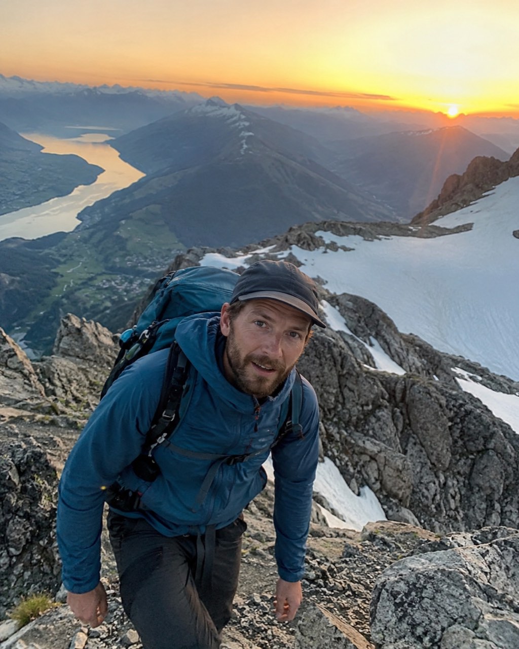 Ausgangsbild: ein Bergsteiger mit blauer Jacke und Rucksack auf einem Grat, im Hintergrund Sonnenaufgang über einem Bergsee
