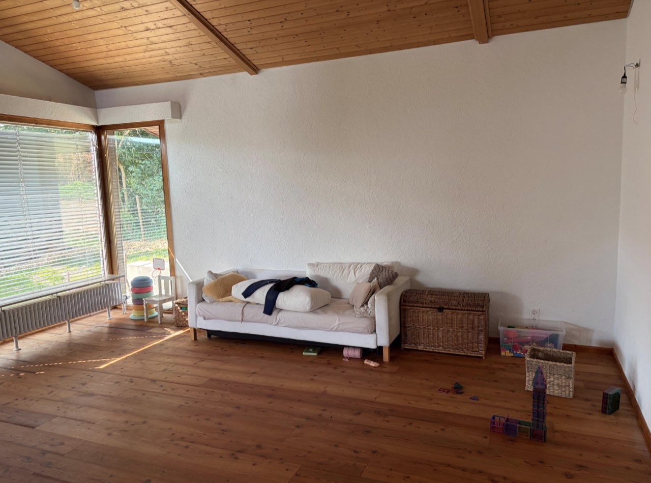 Das Wohnzimmer vorher: weißes Sofa an der Wand, Korbtruhe, Holzdecke, verstreute Kindersachen